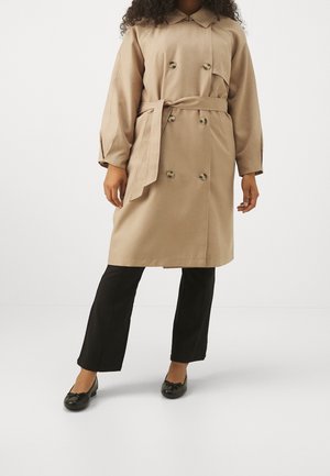 Nainen, jolla on beige kaksirivinen trenssitakki vyötäröhihnalla, mustat polvipituuteen ulottuvat housut ja mustat ballerinat, yksinkertaisen taustan edessä.