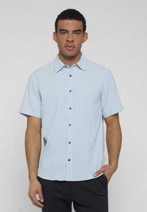 Homme portant une chemise à manches courtes à rayures verticales bleu clair et blanches, avec un pantalon foncé, debout avec une main dans la poche.