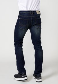 Jeans in denim blu scuro slim-fit con leggera scoloritura, cinque tasche, cuciture a contrasto e una patch sul retro con dettagli del logo. Abbinati a sneaker nere.