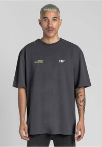 Tmavě šedé oversized tričko vyrobené z bavlny, s malými žlutými a bílými textovými akcenty na přední straně. Krátké rukávy a kulatý výstřih.
