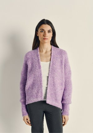 Cardigan in magenta a maglia con un motivo testurizzato, apertura frontale, maniche lunghe e polsini a coste, indossato sopra una maglietta bianca. Design di lunghezza media.