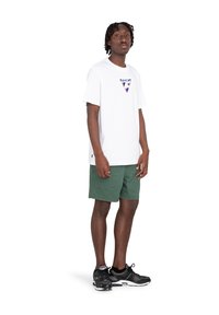 Maglietta bianca in cotone con logo a triangolo multicolore, abbinata a pantaloni corti verdi. Scarpe sportive nere con dettagli in mesh.