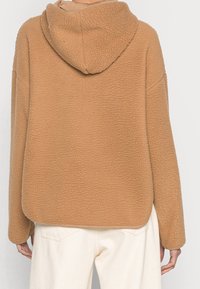 Felpa con cappuccio marrone in pile con superficie texturizzata, un cappuccio e una vestibilità rilassata. L'orlo è arrotondato e viene abbinata a pantaloni di colore chiaro.