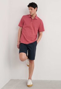 Červené polo triko s krátkým rukávem a třemi knoflíky, spárované s tmavě modrými šortkami a bílými teniskami s béžovými podešvemi.