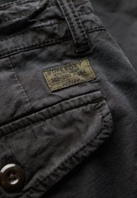 Zwarte stof met een subtiele textuur heeft een zak met een groen label waarop "Superdry Utility Wear" staat. Knopdetail aan de zijkant.