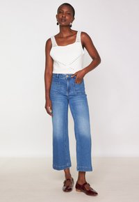 Femme portant un haut en tricot blanc sans manches, un jean bleu taille haute à jambe large avec des poches avant et des chaussures Mary Jane rouge foncé.