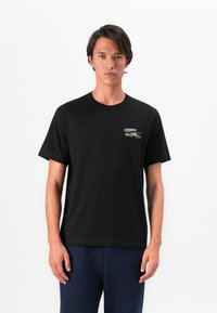 T-shirt en coton noir avec un col rond, des manches courtes et un petit logo brodé sur la poitrine. Texture lisse et coupe classique.