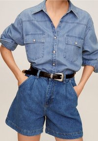 Femme portant une chemise en jean clair à boutons avec les manches retroussées, rentrée dans un short en jean foncé taille haute avec une ceinture noire.
