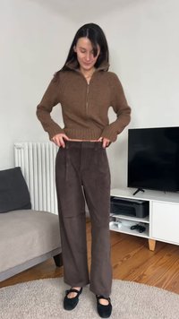 Camisola castanha de malha com gola de fecho éclair combinada com calças castanhas de bombazina de perna larga. Sapatos pretos com detalhe de fivela. Interior claro.