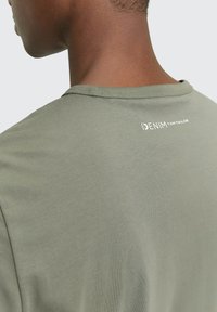 Oliwkowa koszulka z okrągłym dekoltem, o gładkiej fakturze i subtelnym białym logo z tyłu, w pobliżu linii szyi.