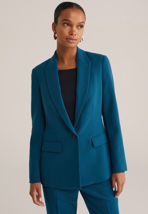 Vrouw in een teal blazer en bijpassende broek met een zwarte top, staande voor een effen lichte achtergrond.