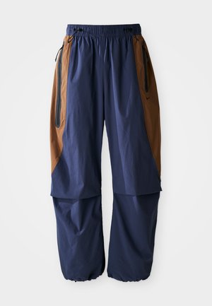 Mørkeblå bukser med brune sidepaneler, elastisk midje og snor. Har to glidelåslommer og et løst, lett design.