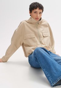 Beige cropped jasje met een hoge kraag, twee voorzakken en een ritsluiting. Gecombineerd met wijde blauwe jeans. Soepele textuur.