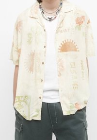 Camicia a maniche corte in tessuto beige chiaro con vari stampati in colori pastello, tra cui fiori e motivi tropicali. Cinque bottoni davanti.
