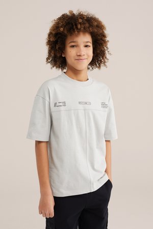 Nooruk käharate juustega, kes kannab heledat halli trükitud T-särki ja musti pükse, seistes ühe käega taskus ühetaustalise tausta ees.