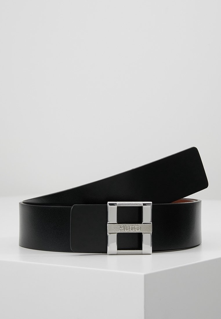 Ceinture en cuir noir avec une boucle rectangulaire en argent gravée de "HUGO", posée sur une surface blanche sur un fond gris clair.
