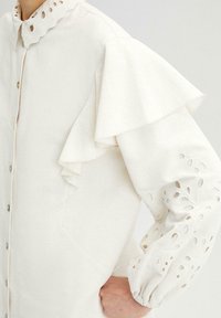 Vitt skjorta med knappar och volangärmar, med broderade utskärningar och en scalloped krage. Tillverkad av ett texturerat tyg.