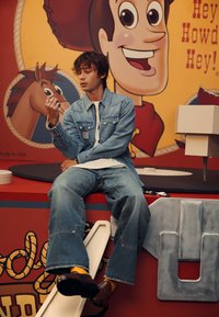 Chaqueta de mezclilla y jeans de pierna ancha en lavado claro, combinados con zapatos marrones y calcetines amarillos. Un personaje de dibujos animados es visible en el fondo.