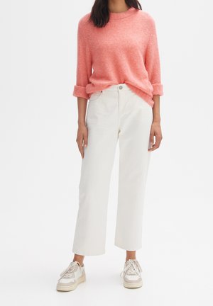 Pull en maille rose avec manches retroussées, associé à un jean blanc à jambes larges et des baskets beiges, sur un fond blanc uni.