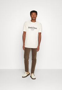 Off-white T-Shirt mit einem "Broken Heart"-Grafik, kombiniert mit braunen Hosen und hellen Sandalen. Schlichtes Design, lockere Passform.