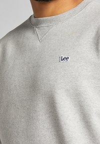 Szara bluza z okrągłym dekoltem, prążkowanymi detalami na kołnierzu oraz małym granatowym logo "Lee" po lewej stronie na piersi. Miękka tekstura.