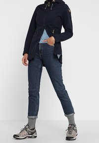 Veste imperméable bleu marine avec fermeture éclair, capuche doublée et cordons de serrage, associée à un haut bleu clair, un jean foncé et des chaussettes grises. Les chaussures sont noires et roses.