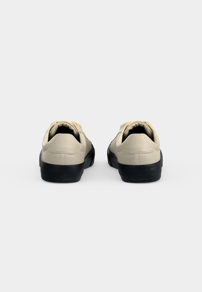 Ζευγάρι μπεζ καμβά sneakers με μαύρες λαστιχένιες σόλες, που απεικονίζονται από πίσω σε απλό ανοιχτό γκρι φόντο.
