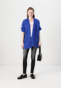 Blått, lodden cardigan med åpen front, brukt over en lys rosa t-skjorte med motiv, sammen med grå skinny jeans og svarte loafer-sko, mens jeg holder en svart veske.