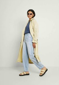 Trench coat bege claro sobre uma blusa azul, combinado com jeans azul claro e sandálias amarelas com sola preta. Os acessórios incluem óculos de sol.