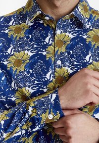 Camisa floral azul con girasoles amarillos, base blanca, cierres de botón, mangas largas y patrones florales intrincados en toda la tela.