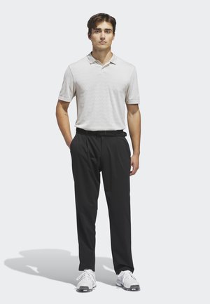 Jonge man staat met handen in de zakken, draagt een lichtgrijze polo, zwarte broek en witte sneakers, tegen een effen lichte achtergrond.