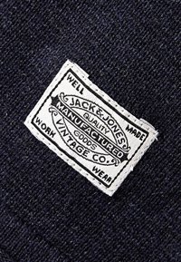 Bílý obdélníkový látkový štítek s černým textem "Jack & Jones Manufactured Goods Vintage Co." přišitý na tmavě modré strukturované pletenině.