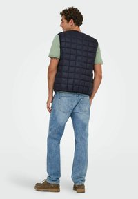 Veste matelassée marine avec une coupe décontractée, dotée d'un col rond et de coutures verticales, associée à un jean bleu clair et des chaussures marron.