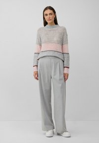 Ženska stoji oblečena v sivi in roza prugasti pulover, svetlosivih širokih hlačah, belih športnih copatih in srebrnih obročastih uhanskih. Ozadje je enostavno.