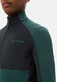 Veste zippée verte et noire avec col montant, fabriquée en tissu lisse. Présente un logo Vaude sur la poitrine.