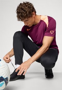 Roupa desportiva: camisola de manga curta cor vinho com detalhes laranja, calças pretas, sapatilhas de futebol pretas; pessoa a atar os atacadores perto de uma bola de futebol.