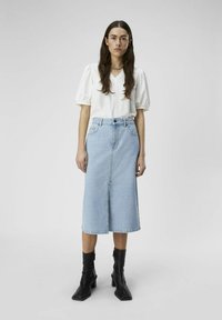 Sai de denim azul claro con abertura frontal, combinado con una blusa blanca de manga corta con escote en V y mangas abullonadas, llevada con botas negras hasta el tobillo.