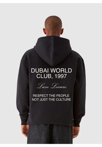 Juodas džemperis su dideliu gobtuvu, ant nugaros su baltu tekstu: "DUBAI WORLD CLUB, 1997 Luxe Leisure GERBKITE ŽMONES, O NE TIK KULTŪRĄ."
