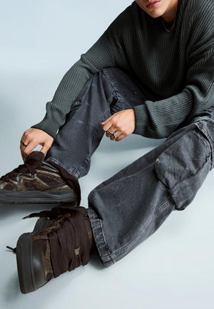 Személy szénszürke cargo nadrágot, sötétbarna, vastag fűzőjű, masszív tornacipőt és sötét bordázott pulóvert viselve ül a padlón, és egy cipőt tart a kezében.