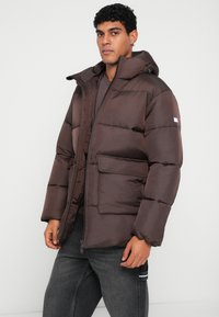 Καφέ τζάκετ puffer με κουκούλα, με φερμουάρ, παχιές ραμμένες περιοχές, μπροστινή τσέπη και λογότυπο στην αριστερή μανσέτα.