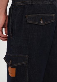 Shorts din denim închis la culoare, cu cusături portocalii, având multiple buzunare, o etichetă din piele cu marcă și un design relaxat.