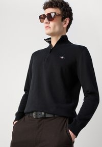 Crni džemper na patentni zatvarač s rebrastom teksturom i malim logotipom. Uparen s tamno smeđim hlačama i velikim kvadratnim sunčanim naočalama smeđe nijanse.