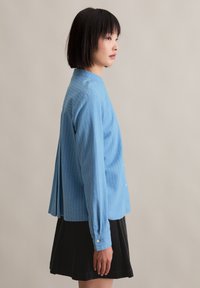 Blauw gestreepte shirt gemaakt van lichtgewicht stof, met een knoopsluiting aan de voorkant, lange mouwen en een geplooide achterkant, gedragen met een zwarte rok.