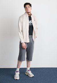 Camicia button-up beige chiaro sopra una maglietta nera con logo, pantaloni corti grigi e calze bianche con scarpe da ginnastica con dettagli viola. Sfondo semplice.