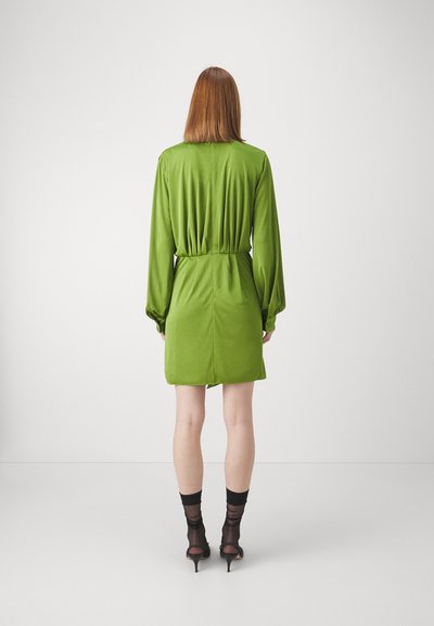 Rochie verde din satin cu mâneci lungi și talie strânsă, având o textură fină și o lungime scurtă. Combinată cu șosete negre transparente și pantofi cu toc.
