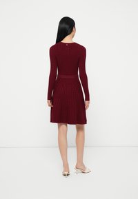 Donna con lunghi capelli neri che indossa un vestito di maglia bordeaux con cintura e mule bianchi con tacco, in piedi contro uno sfondo bianco semplice.