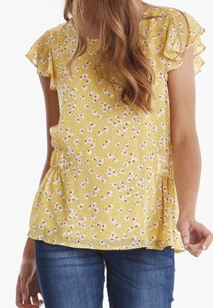 Jeune femme portant un chemisier jaune à manches courtes à imprimé floral avec des épaules volantées et un jean bleu, debout sur un fond blanc.