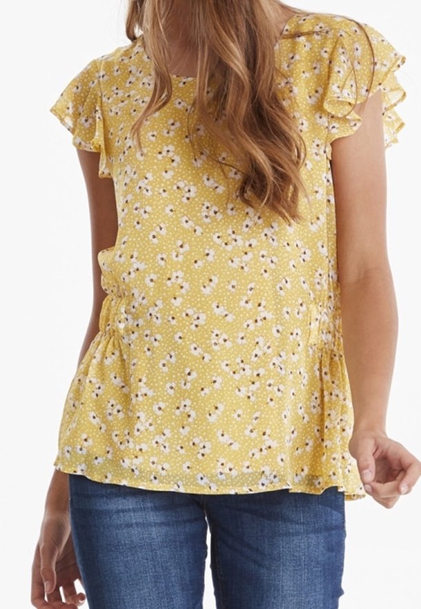 Jeune femme portant un chemisier jaune à manches courtes à imprimé floral avec des épaules volantées et un jean bleu, debout sur un fond blanc.