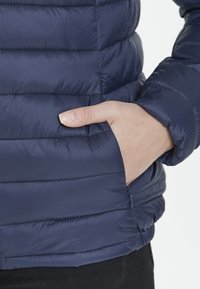 Marinblå puffjacka med horisontella kviltade sektioner, med en sidoficka och elastiska ärmslut. Slät, lättviktsmaterial med en liten glans.