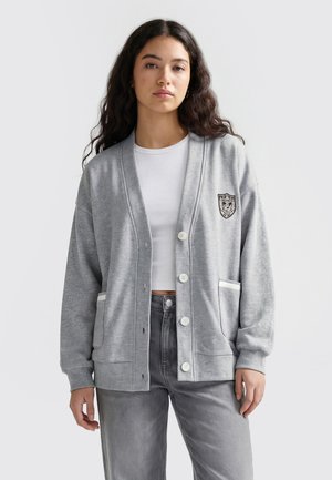Jeune femme aux longs cheveux foncés portant un cardigan gris clair boutonné par-dessus un crop top blanc et un jean gris, debout devant un fond uni.